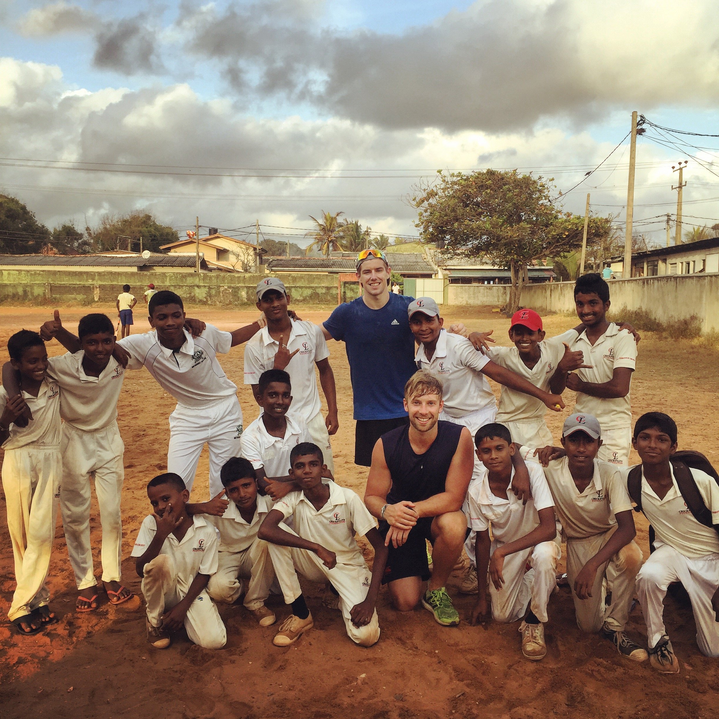 A group of children and two adults posing on a dirt field, with clouds overhead and houses in the background. The children are wearing sports uniforms and the adults are casually dressed.