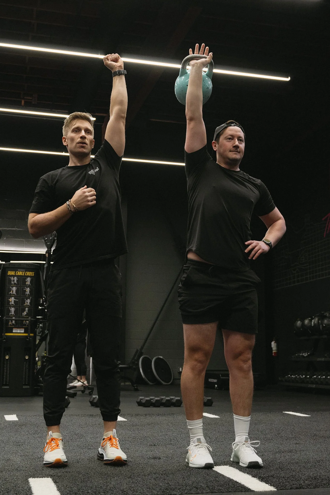 Two men in athletic clothing stand in a gym, one holding a kettlebell overhead, the other with a fist raised. Both wear black t-shirts, black shorts, and sneakers. The gym has black walls, rubber flooring, and gym equipment in the background.