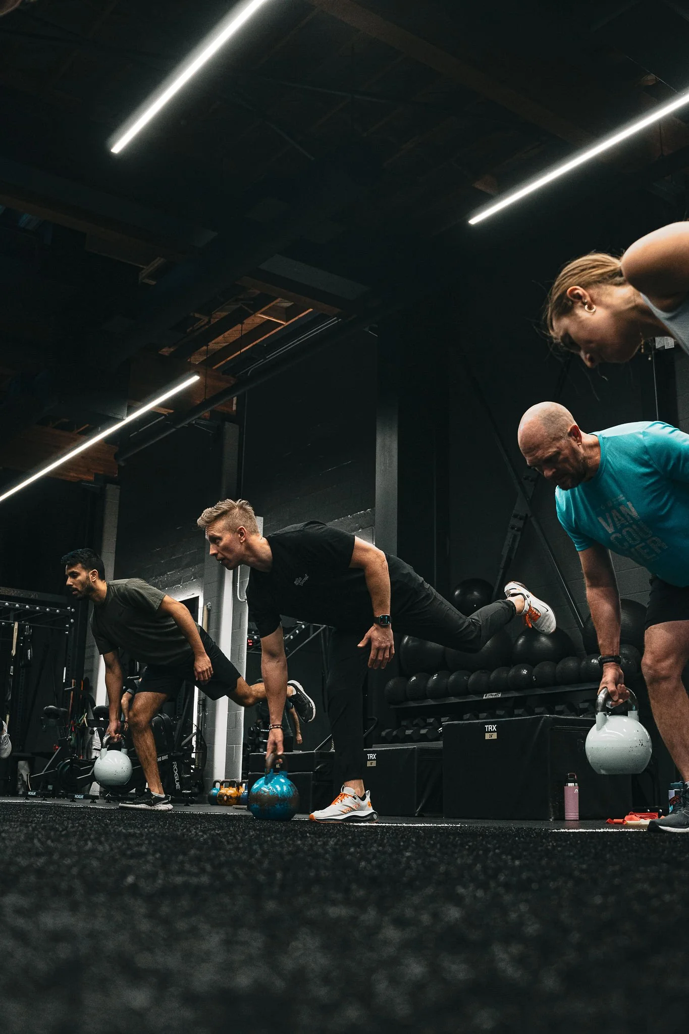 Group of people performing kettlebell swings during a workout session in a dark gym with ceiling lights and black workout equipment.