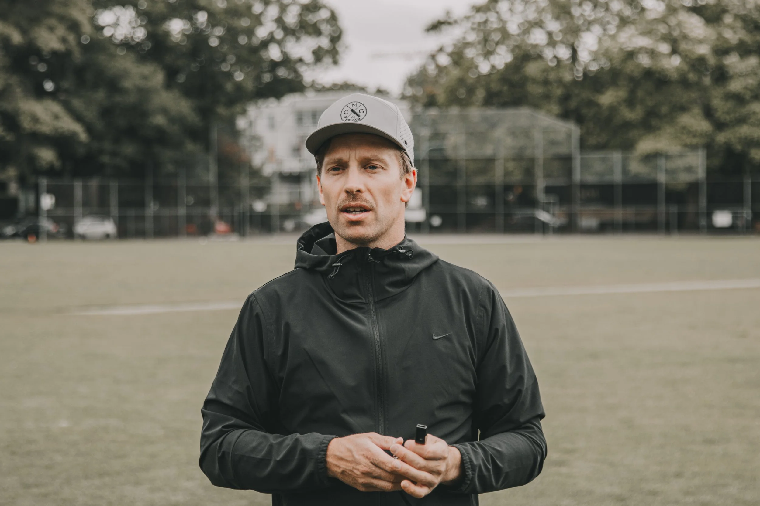 A man dressed in a black Nike jacket and a gray baseball cap stands outdoors on a field, holding a small object in his hands, with a tennis court and cars in the background.