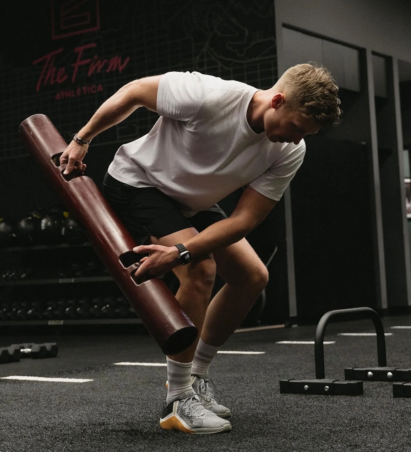 A young man in a gym performing a workout with a cylindrical foam roller, wearing a white t-shirt, black shorts, and sneakers.