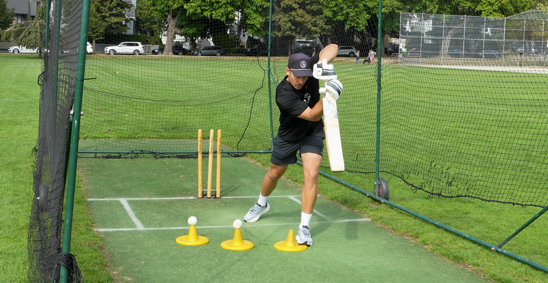 Cricketer practicing batting drills in cricket practice net on grass field, with batting pads, stumps, and cones.
