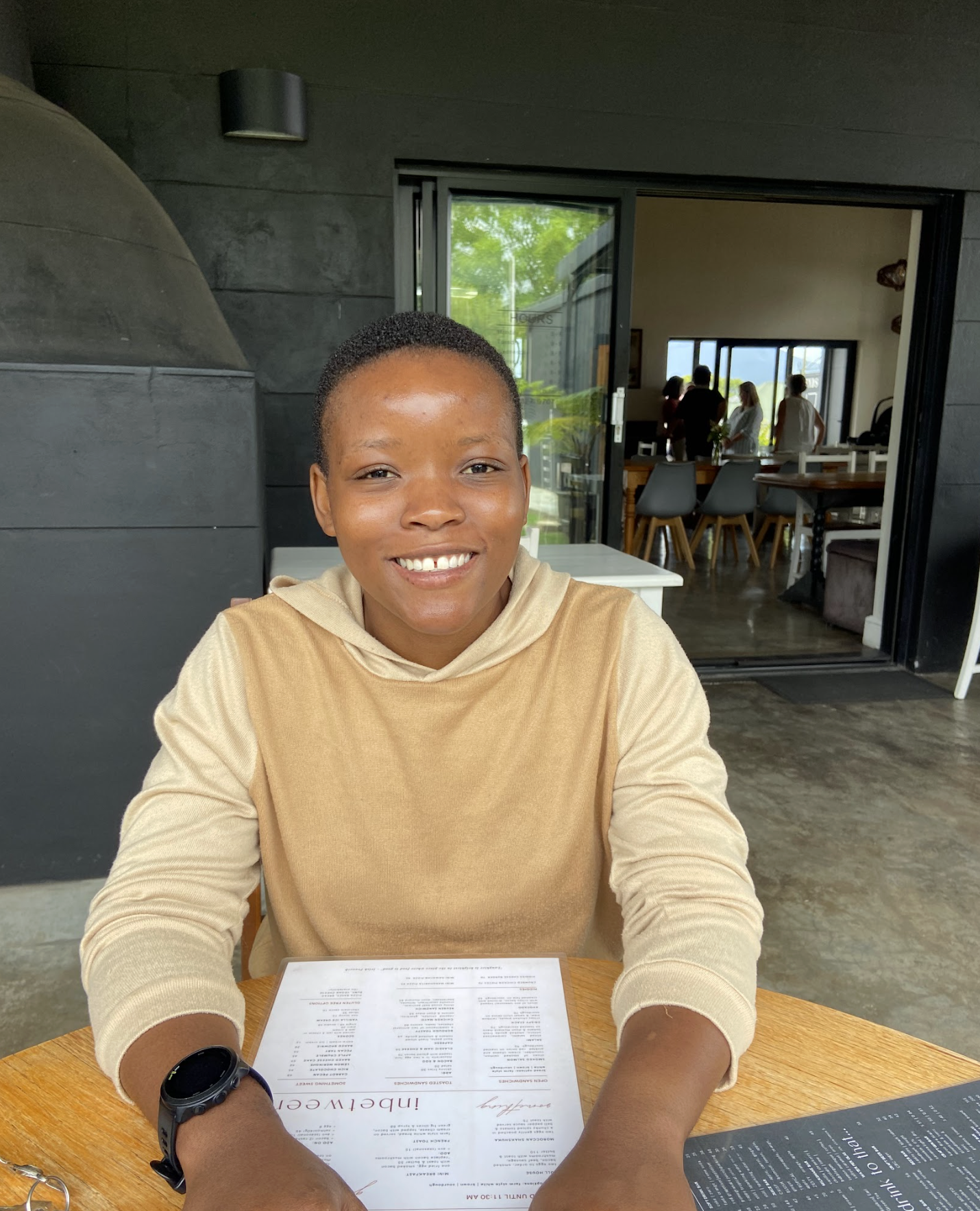 A smiling woman with short hair, sitting at a wooden table in a restaurant or cafe, with a menu in front of her. Behind her, a glass door reveals other people inside the establishment, and the decor is modern and minimalist.