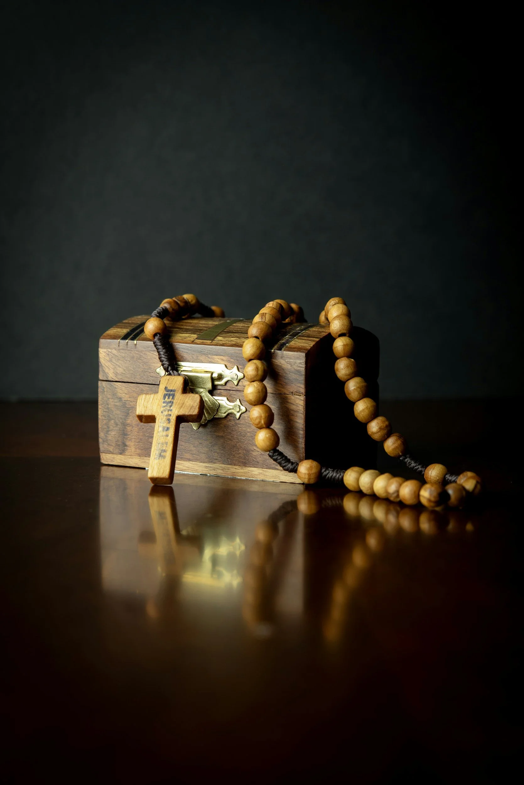 A small wooden box with a latch, positioned on a reflective surface against a dark background. A string of wooden prayer beads and a wooden cross tag are draped over the box. The cross tag has etched writing on it.