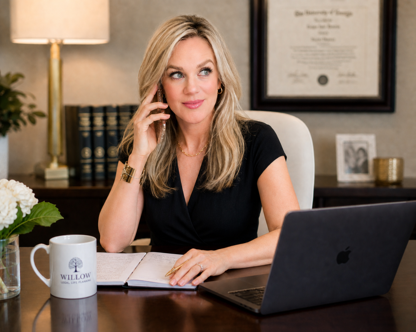 A woman with blonde hair sitting at a desk, talking on the phone. There is a laptop, a notebook, a pen, a mug with the logo 'Willow Legal Life Planning,' and a plant with white flowers on the desk. In the background, there is a framed diploma on the wall, a table lamp, and books.