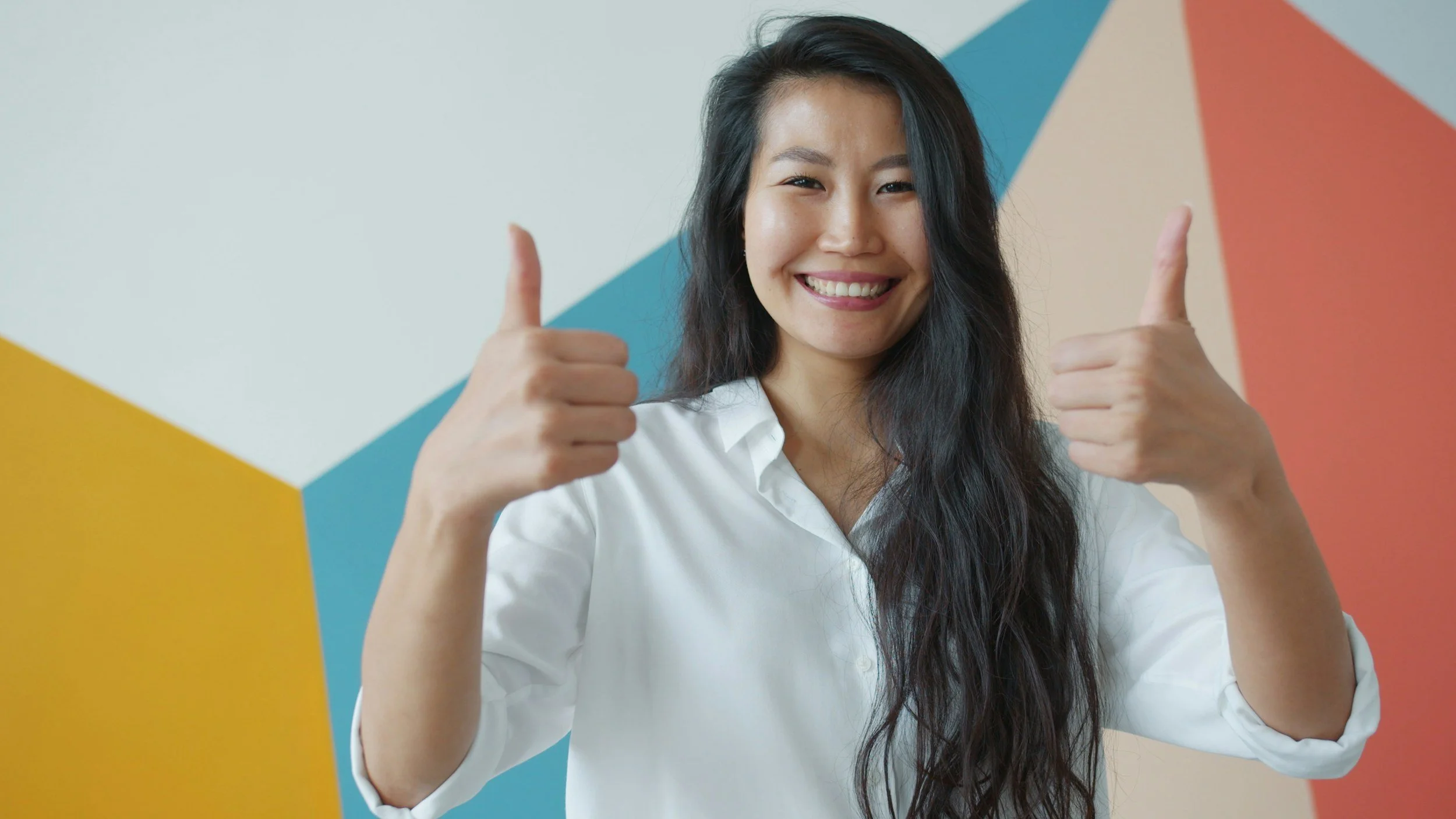A smiling woman with long dark hair wearing a white shirt giving two thumbs up against a colorful geometric background.