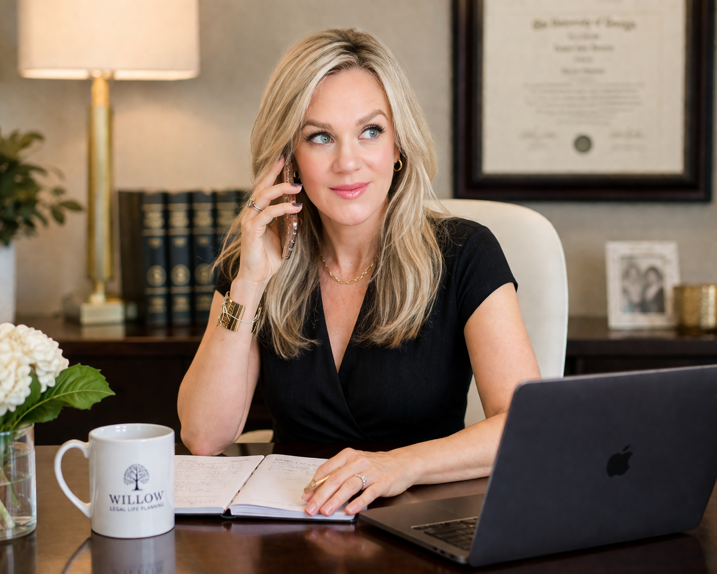 A blonde woman in a black dress sitting at a desk having a phone conversation. She is holding a phone to her ear with her right hand, and her left hand is resting on an open notebook on the desk. She is smiling slightly and looking off to the right. There is a laptop, a white mug with the logo 'WILLOW LEGAL LIFE PLANNING,' a vase of white flowers, a framed photograph, and a picture frame in the background with a diploma on the wall.