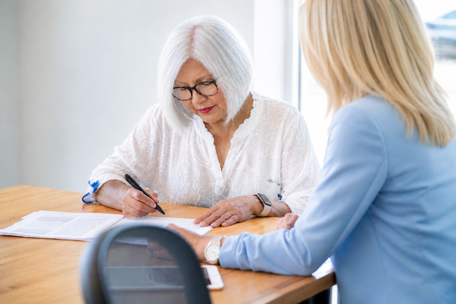 Two women sitting at a wooden table, reviewing documents. One woman with white hair and glasses is signing a paper, while the other woman, with blonde hair, looks on.