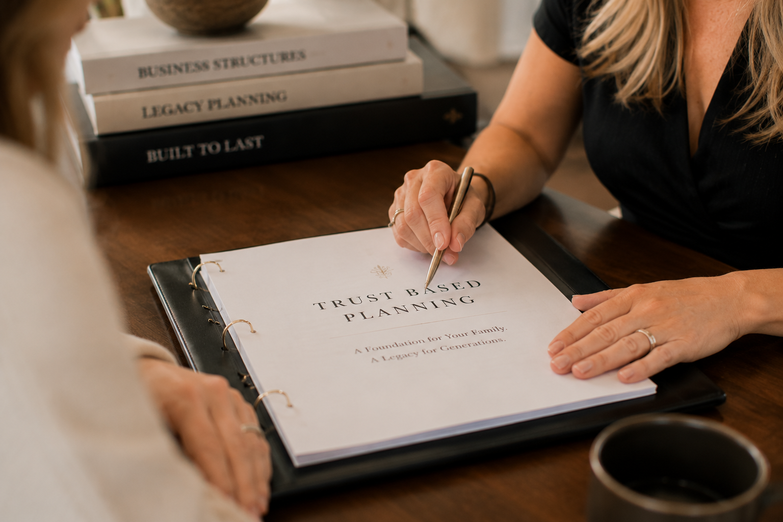 Two women discussing a document titled 'Trust Based Planning' at a wooden table, with a stacked set of books related to business planning in the background.