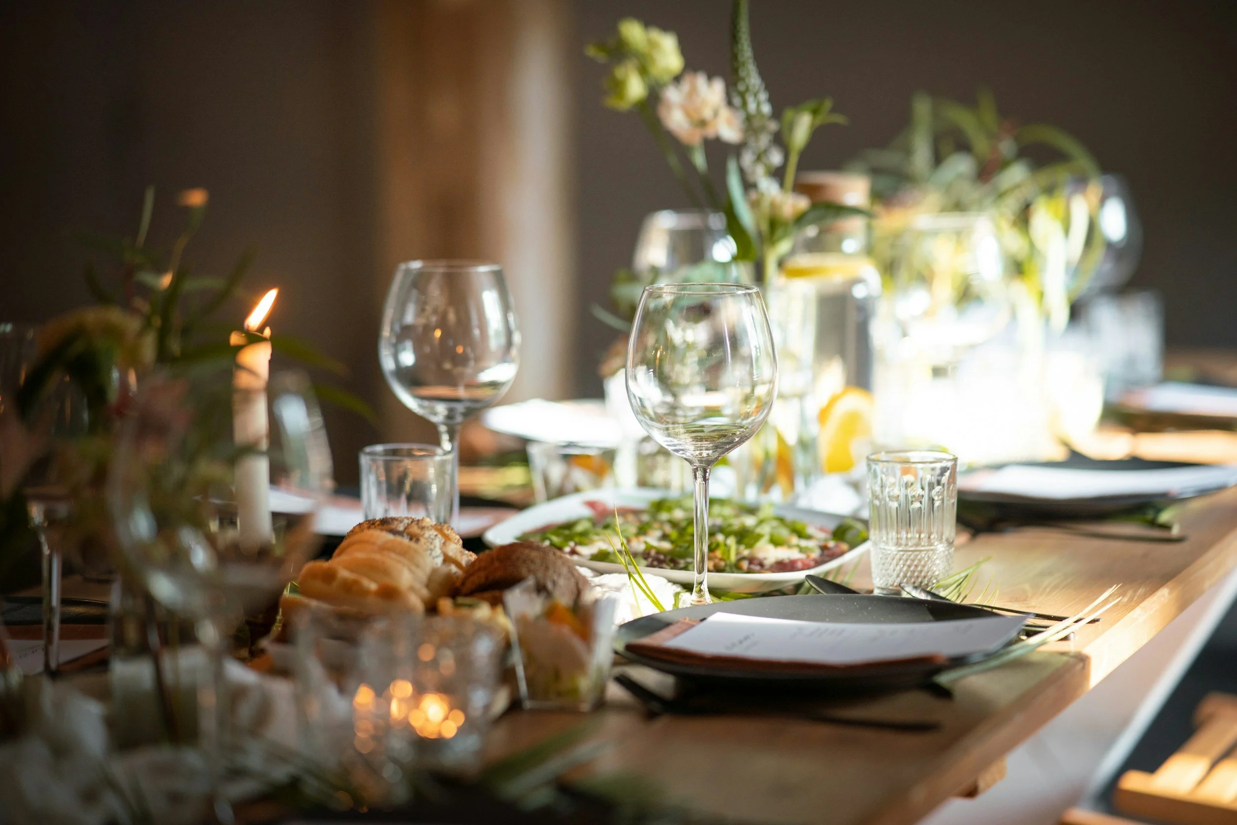 A dining table set for a meal with glassware, flowers, and food including bread and salad, illuminated by natural light.