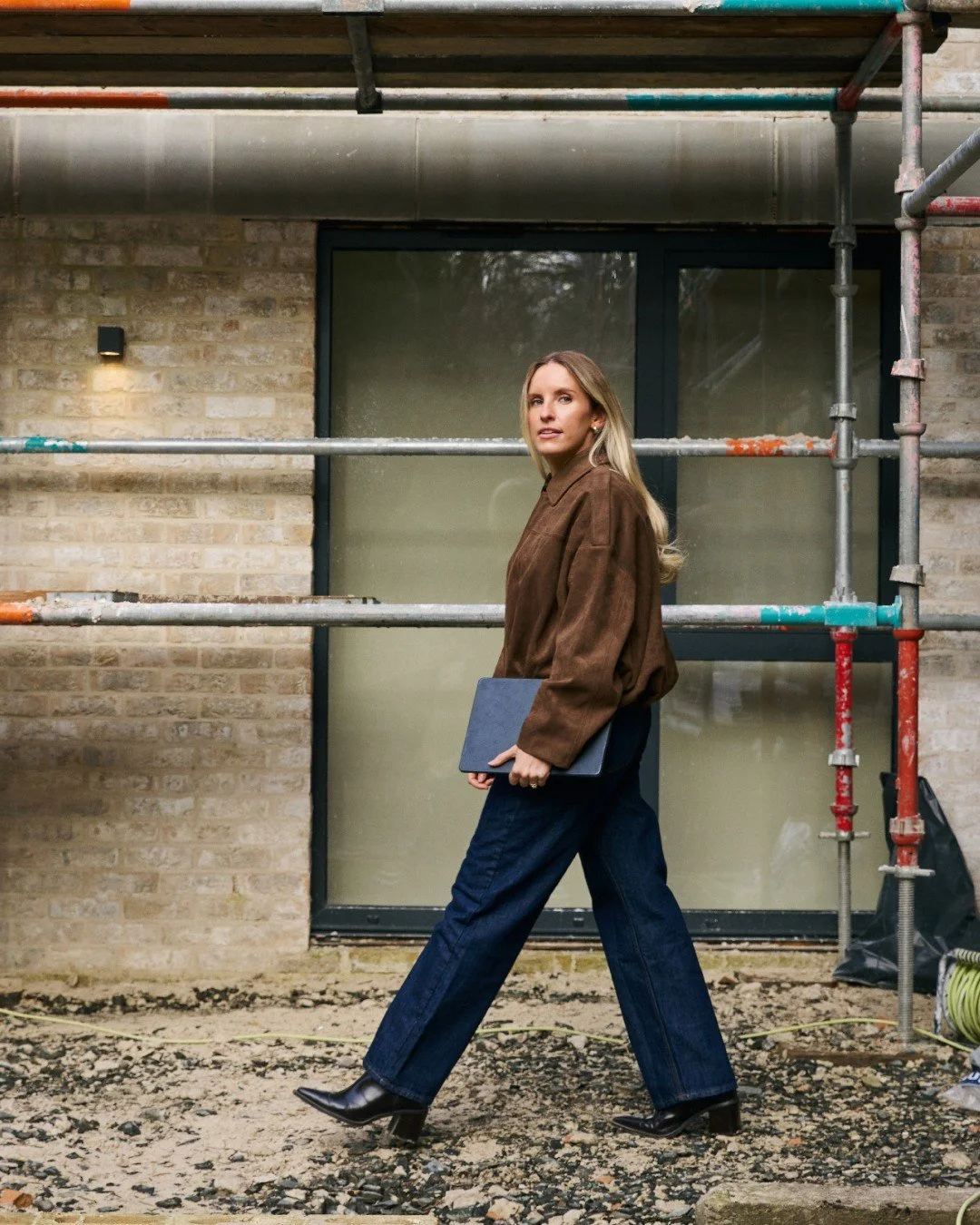 A woman with blonde hair in a brown jacket, dark blue jeans, and black boots walks past a building under construction, holding a closed laptop.