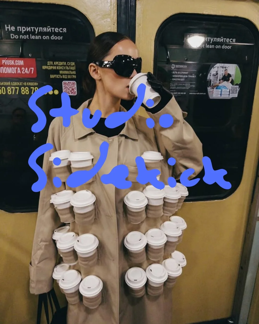 A woman wearing sunglasses and a beige trench coat standing in front of a yellow train door, drinking coffee from a disposable cup. Several more cups are attached to her coat like an accessory. The words 'Stupid girl' are written in blue over the image.