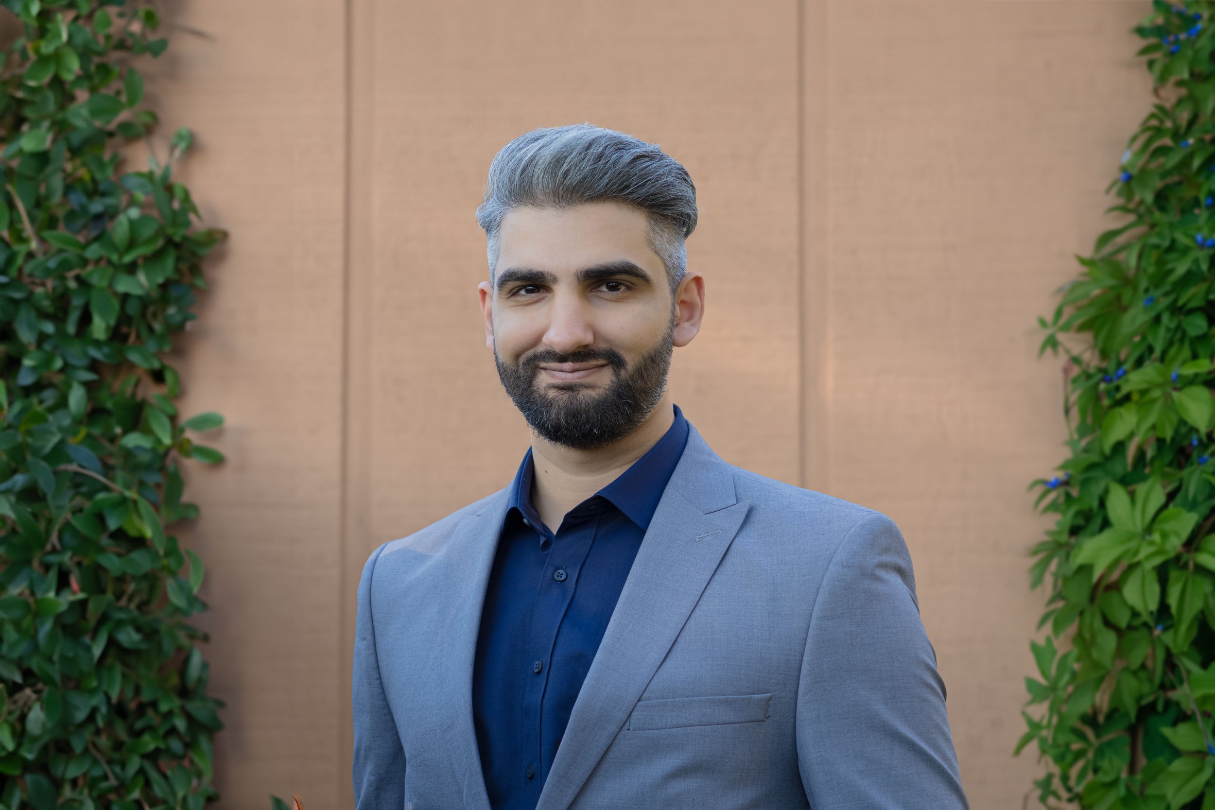 A man with gray hair and a beard, wearing a gray blazer over a dark blue shirt, standing outdoors in front of a wooden wall with green foliage on both sides.