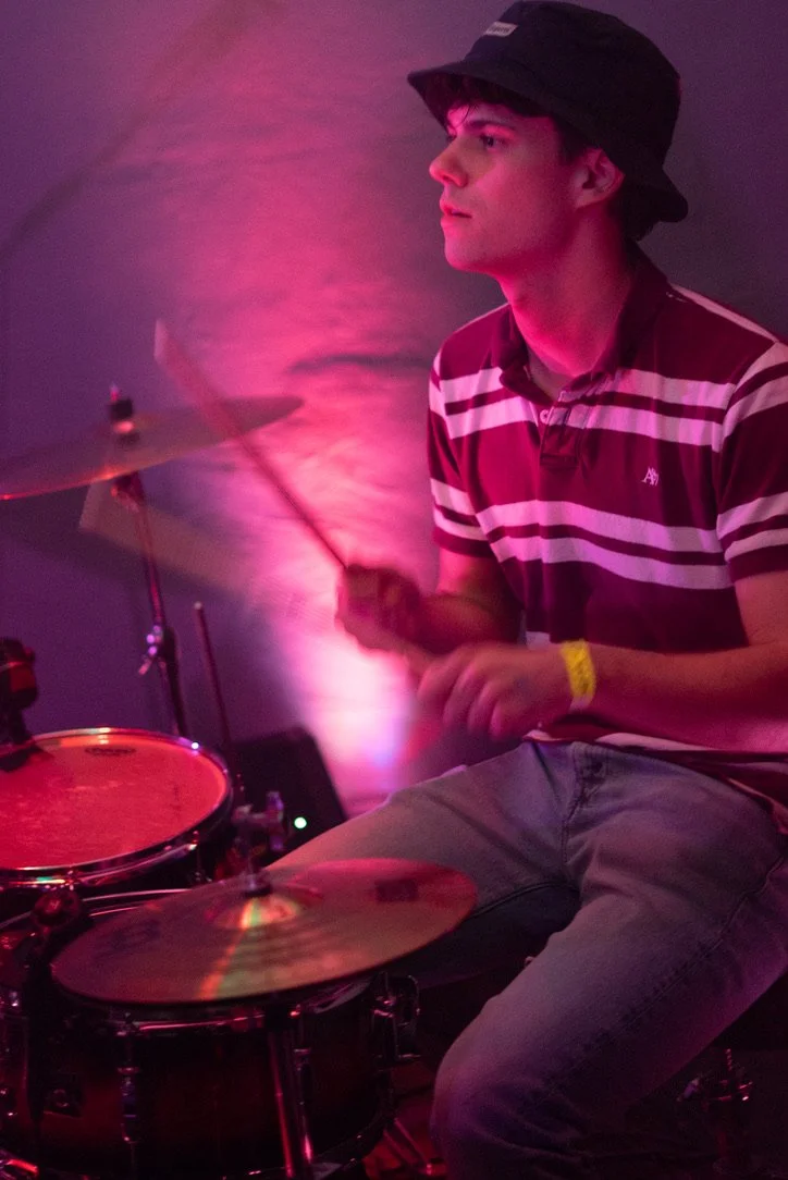 Young man playing drums in a dimly lit room with pink and purple lighting, wearing a striped polo shirt, black bucket hat, and yellow wristband.