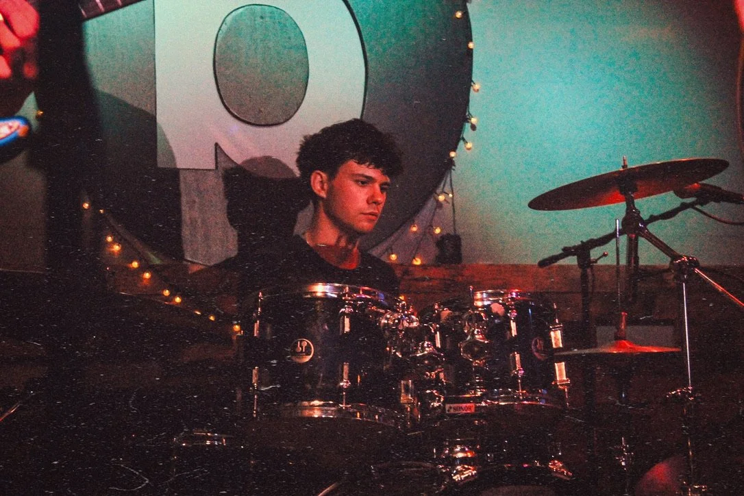 Young man playing the drums on a stage with a colorful background and string lights.