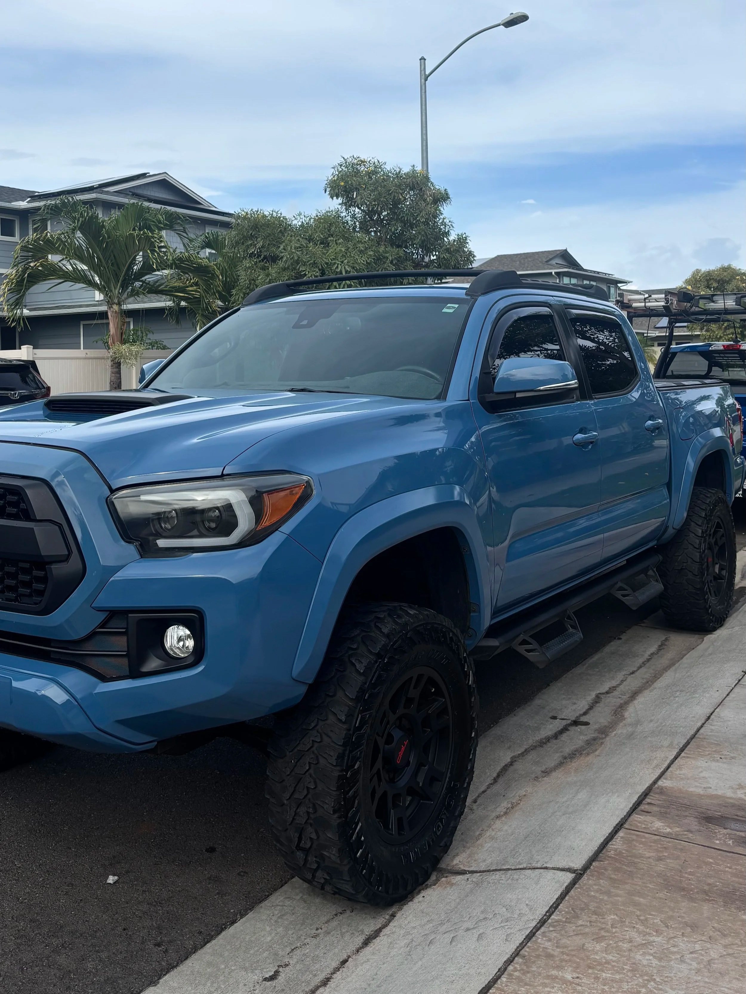 Blue pickup truck parked on the side of a street in a residential neighborhood, with houses, trees, and a streetlamp in the background.