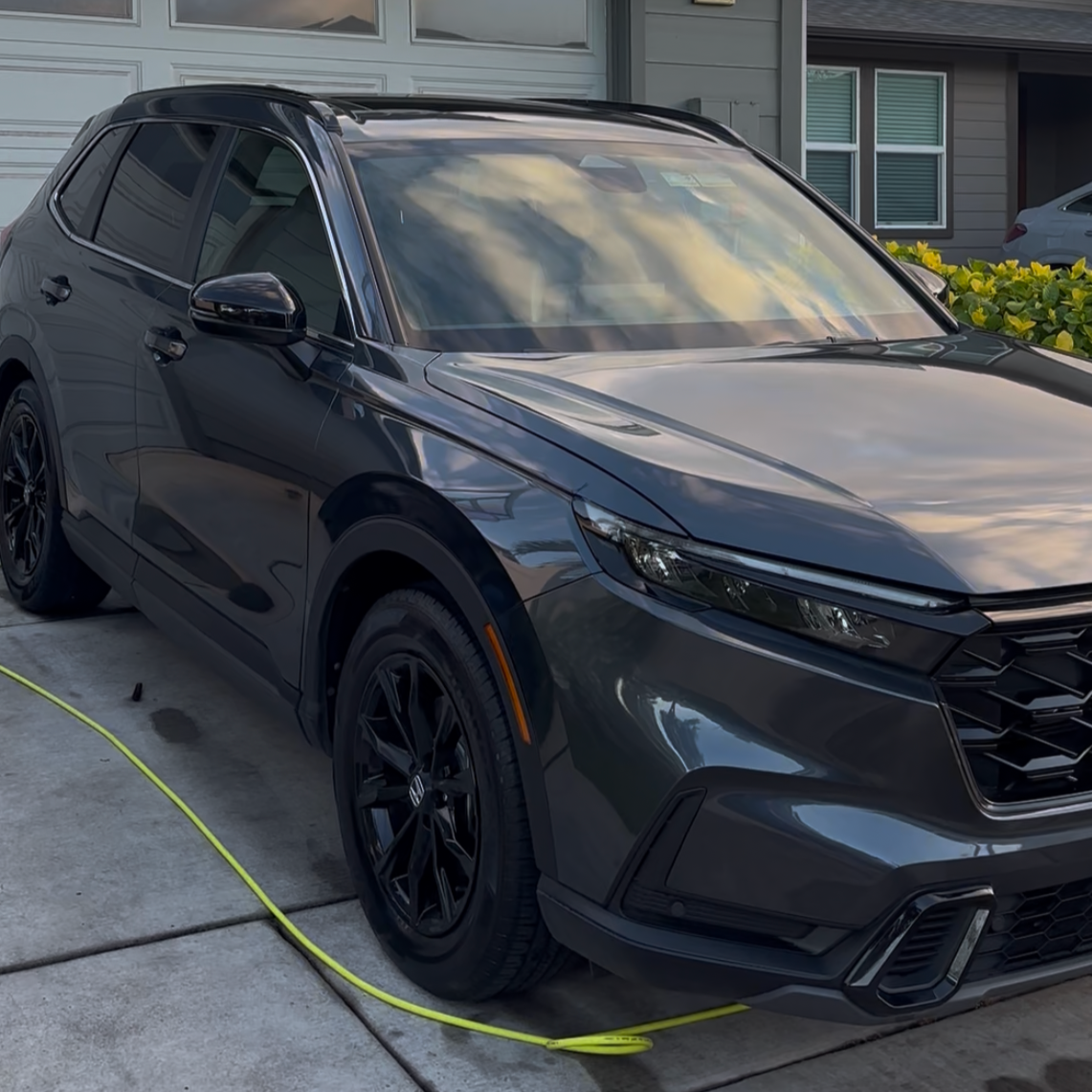 A dark gray or black modern sedan parked on a driveway in front of a house, with a yellow hose on the ground nearby.