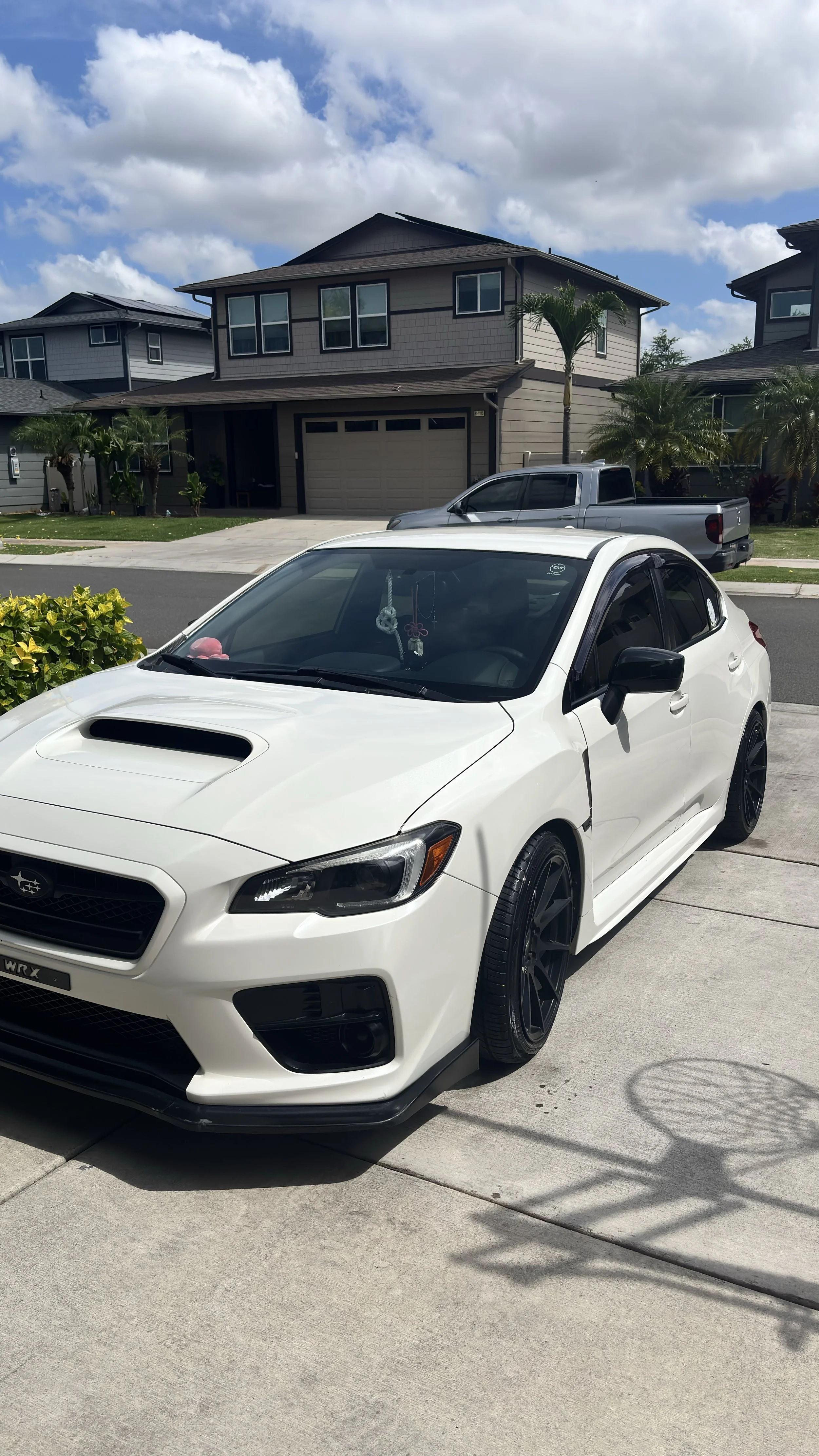 A white Subaru WRX parked on a driveway in front of a modern two-story house with palm trees and a blue sky with scattered clouds.