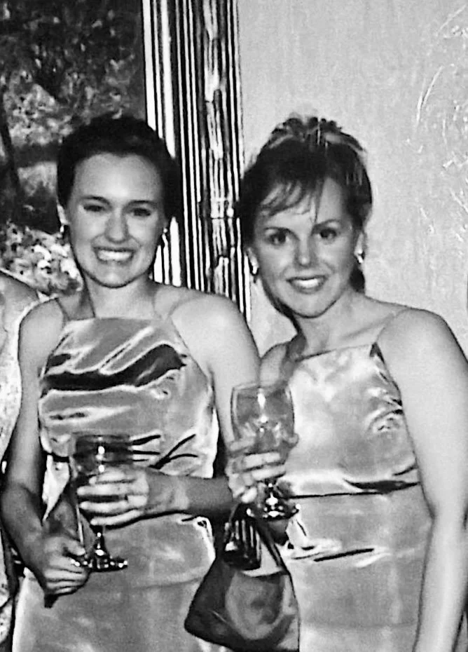 Two women in elegant satin dresses smiling and holding wine glasses at a social gathering or party.