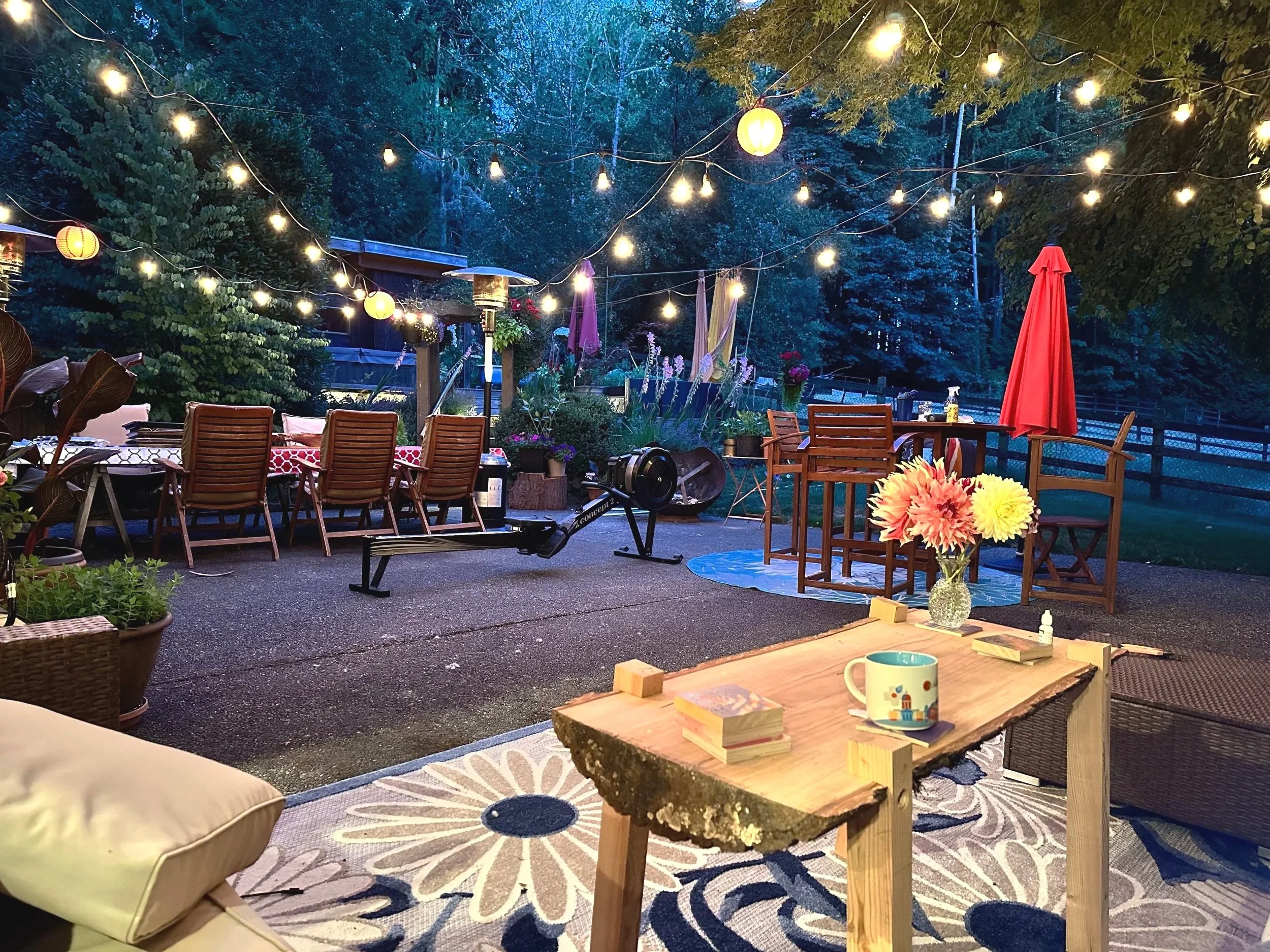 An outdoor evening patio scene decorated with string lights and lanterns hanging above. There are wooden chairs and tables with umbrellas, potted plants, and flowers. A treadmill is positioned in the yard and a beverage mug and game pieces on a rustic wooden table in the foreground. There is trees and a fence in the background.