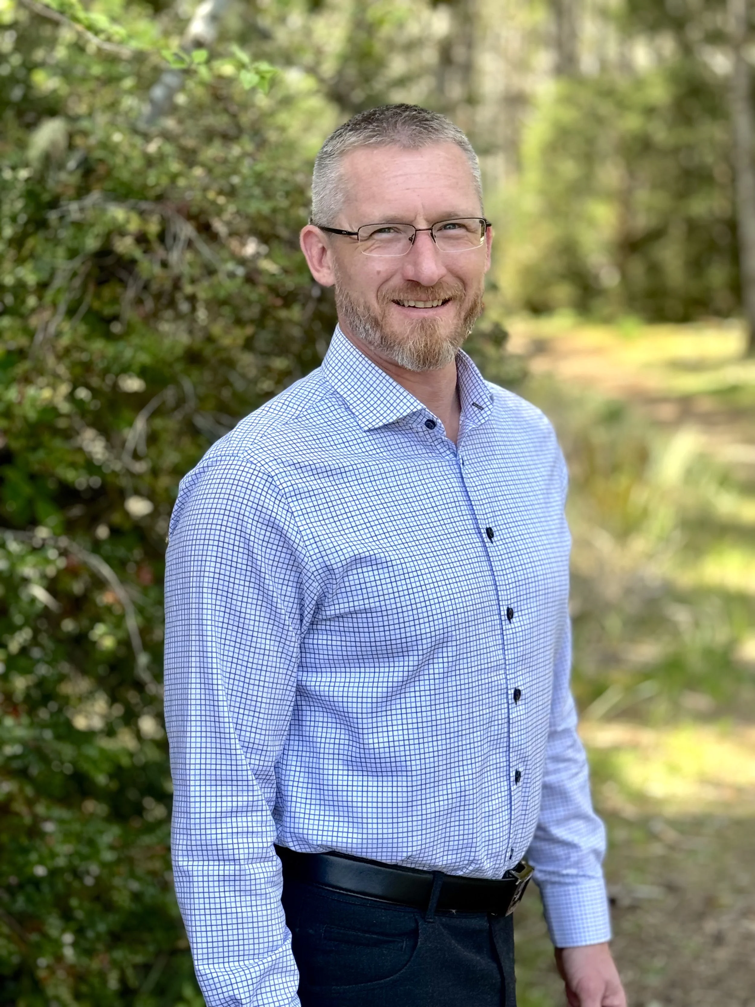 Edward Welter who is running for Sheriff of Grays Harbor is standing outdoors in Grays Harbor County among green foliage and trees.