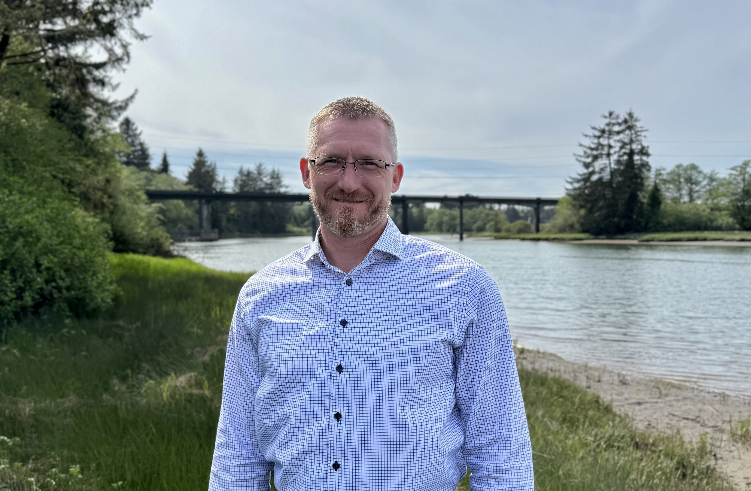 Ed Welter, candidate for Grays Harbor Sheriff, is smiling in front of John's River in Grays Harbor County, with trees, a bridge, and a cloudy sky in the background.
