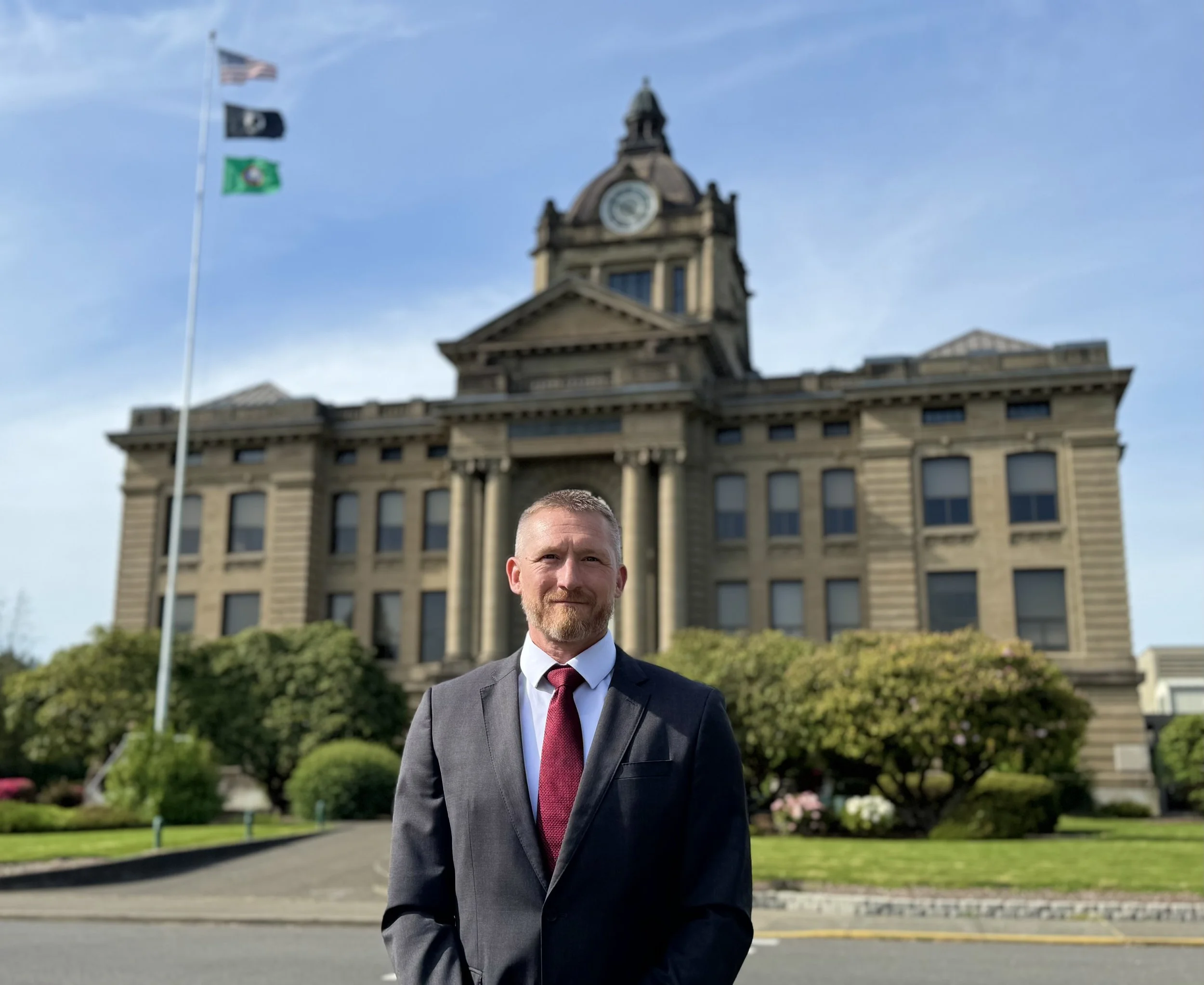 Edward Welter in a suit and red tie standing outdoors in front of Grays Harbor County Courthouse with flags, and a statue, with trees and flowers in the background.
