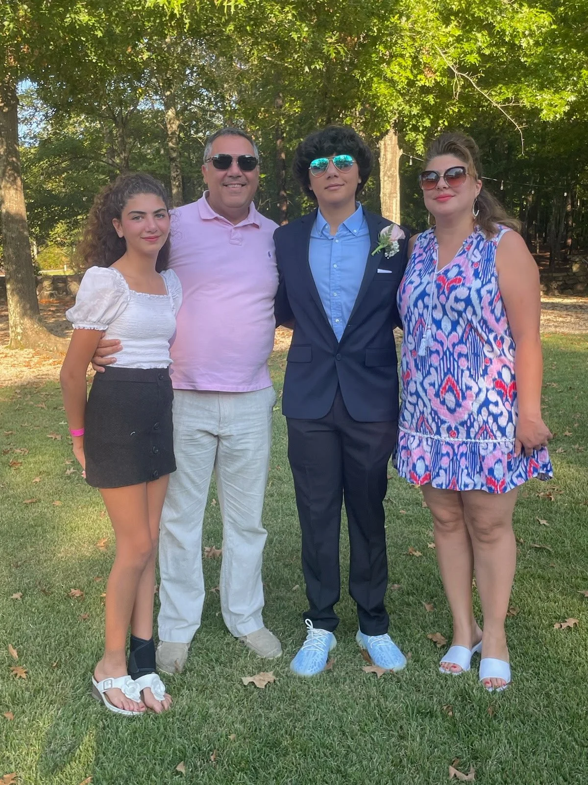 Four people standing outdoors on grass with trees in the background. Two women on the left and right, a man and a person in a suit in the middle. All are smiling and wearing sunglasses, dressed casually or semi-formally, with the person in a suit wearing a boutonniere.