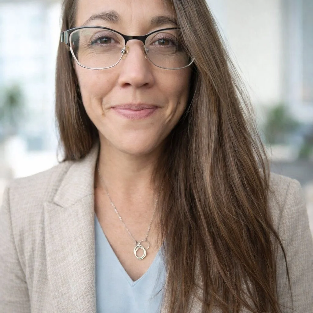 A woman with long brown hair wearing glasses, a beige blazer, and a silver necklace, smiling softly.