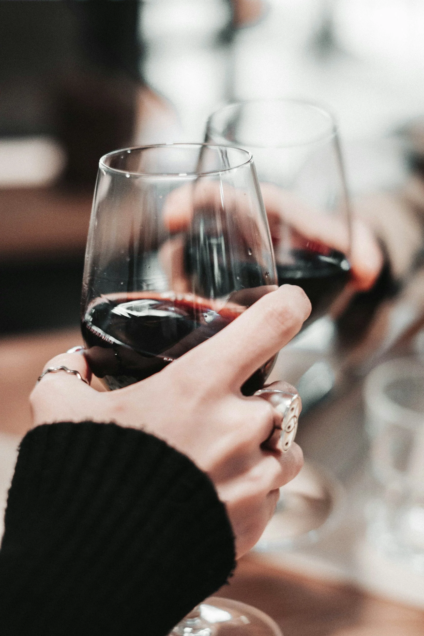 Close-up of a hand holding a glass of red wine, with other glasses in the background, in a social setting.