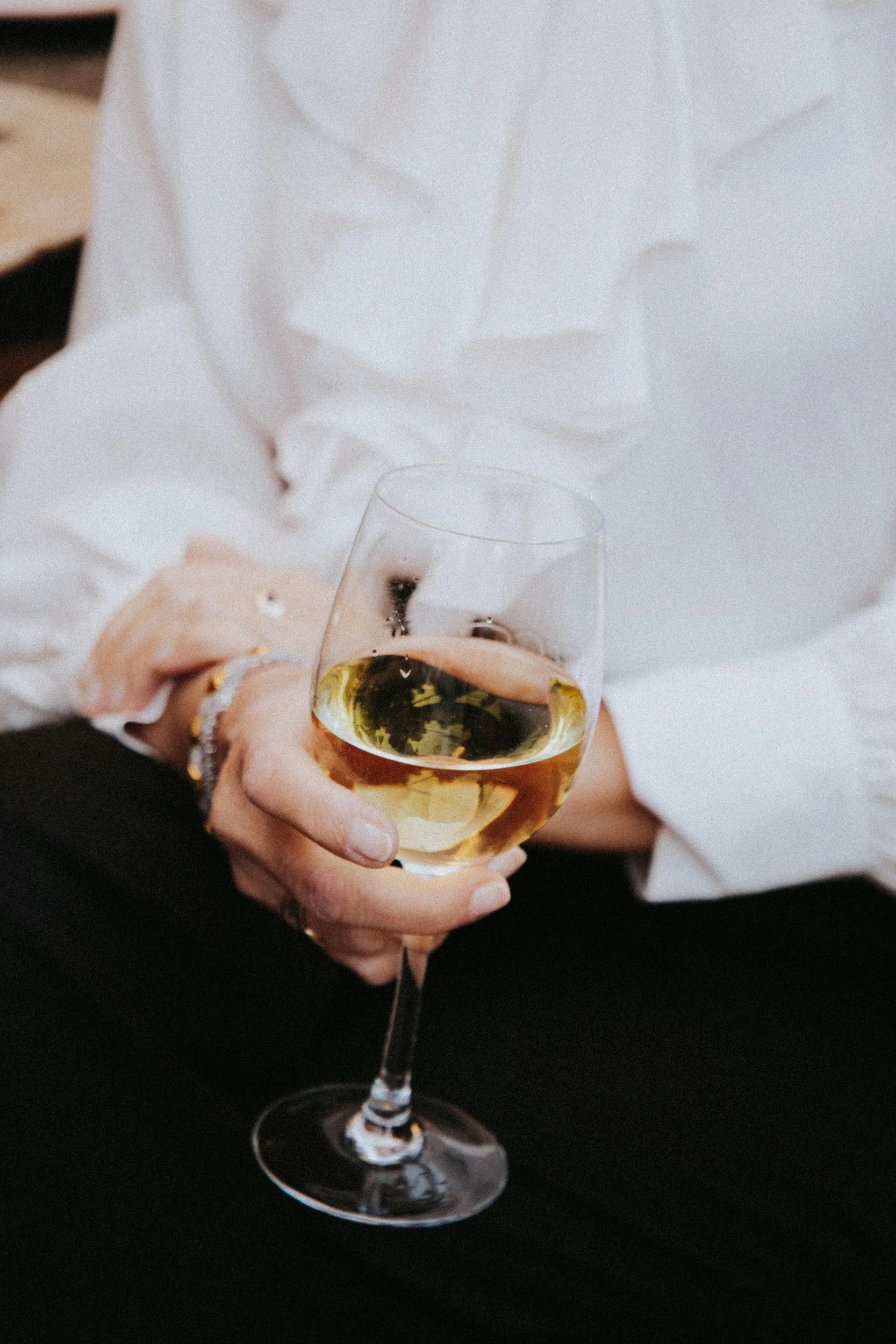 Person in a white shirt holding a glass of white wine, with jewelry on their fingers and wrist, against a dark background.