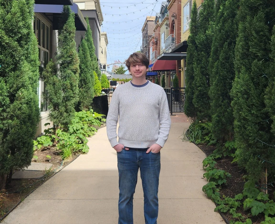 A young man with brown hair wearing a gray sweater and blue jeans standing on a sidewalk between green bushes and buildings.