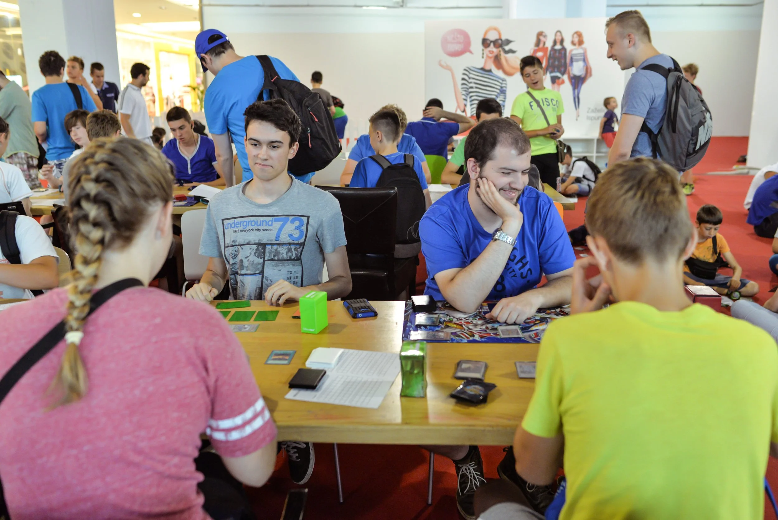 A group of teenagers and young people playing card games at a large indoor event or gathering, with some sitting at tables and others standing around.