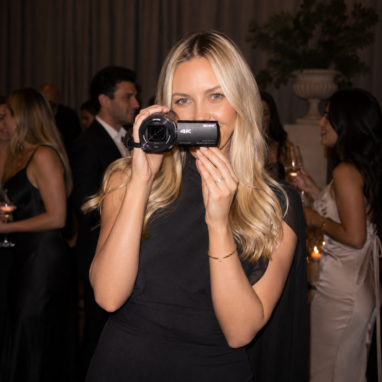 A blonde woman in a black dress holding a 4K camcorder rental from film yours at a formal event with other guests in the background.