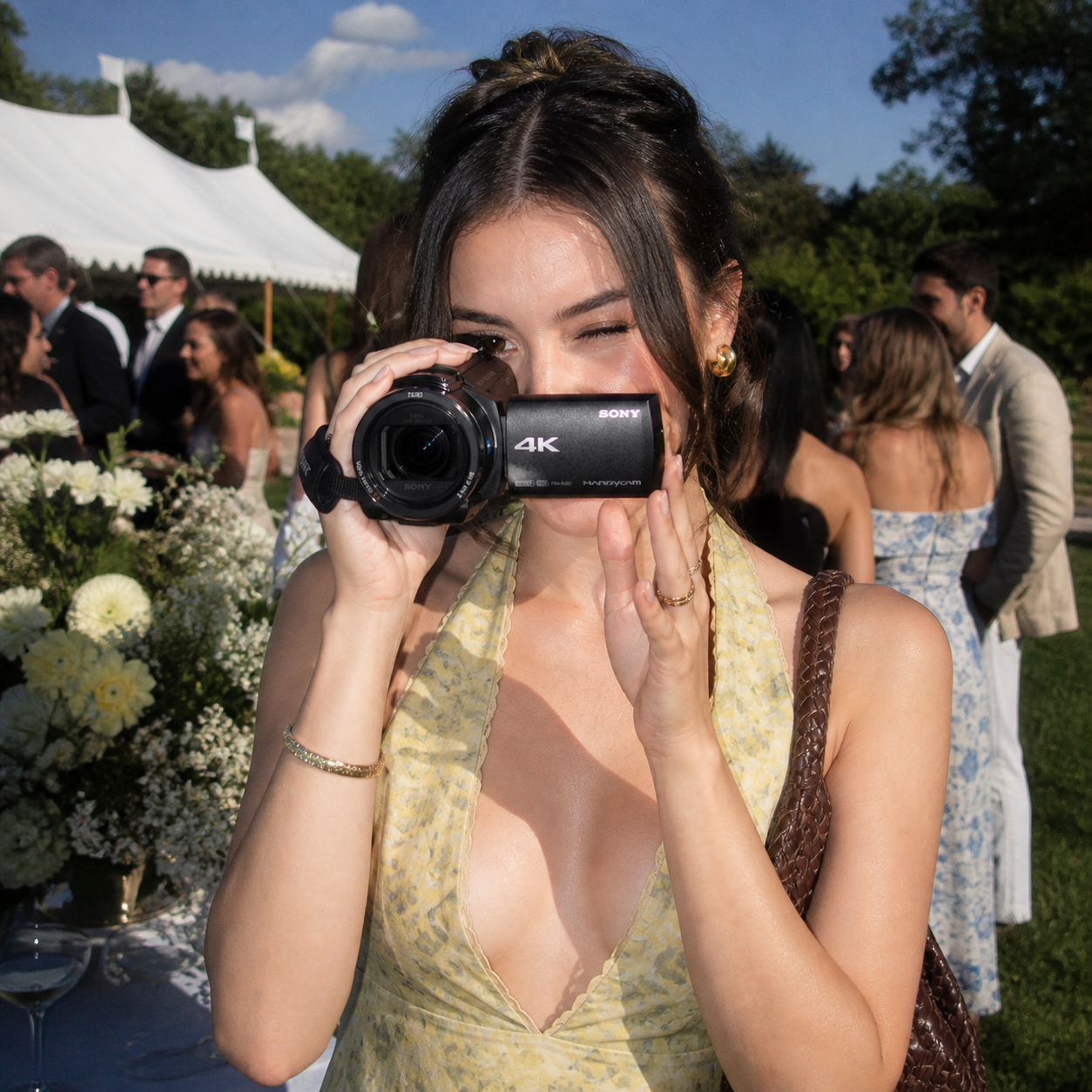 Young woman in yellow dress filming with a 4K Sony camcorder at an outdoor wedding reception, with guests socializing and white tents in the background.