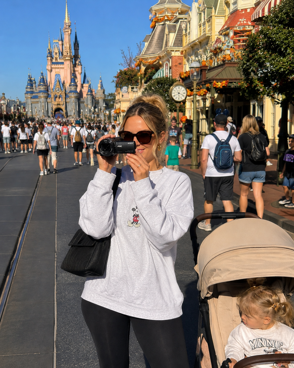 A woman with sunglasses using our camcorder rental at Disneyland, with the castle in the background and a stroller with a young girl nearby capturing her family vacation.