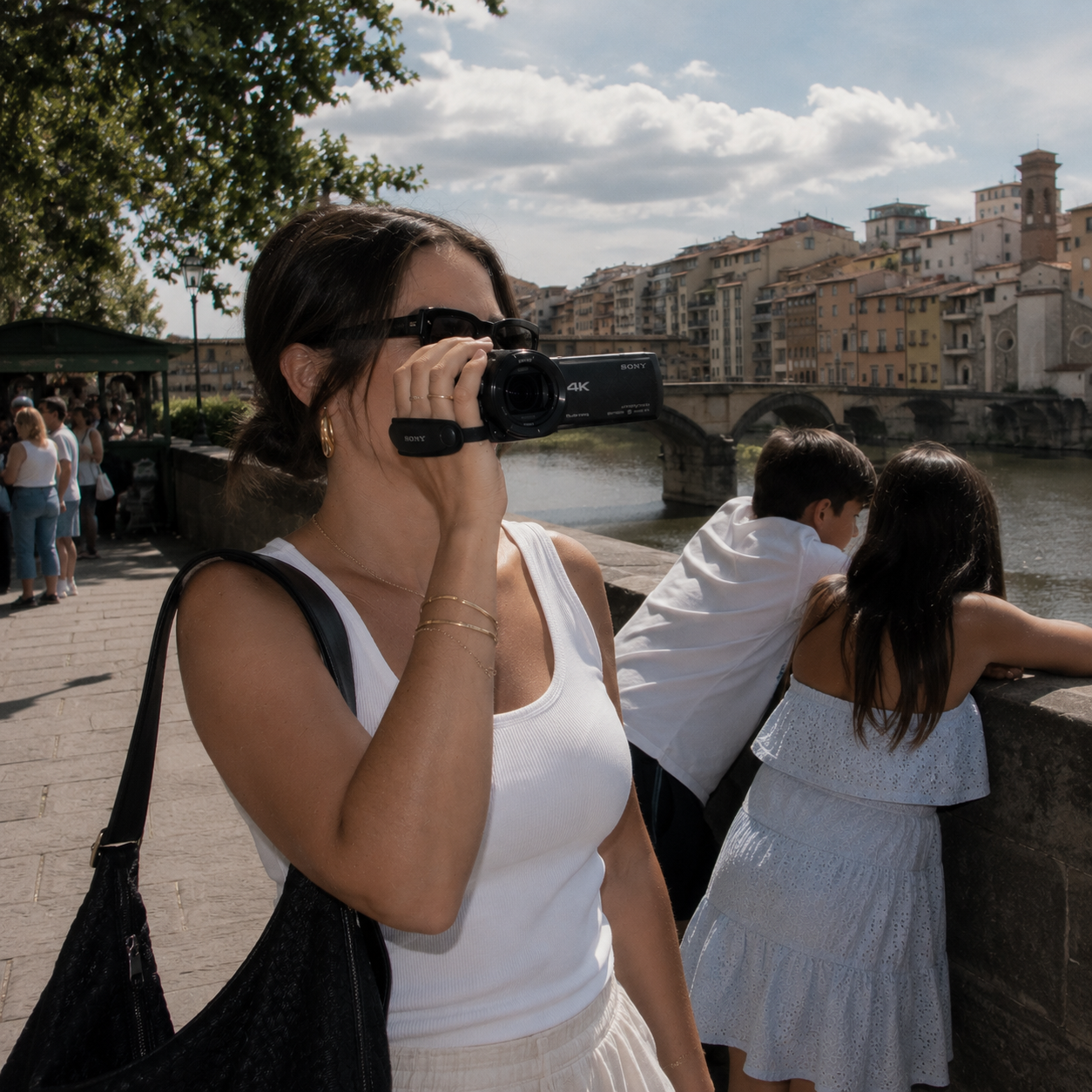 A woman taking a video with a camcorder rental by the river with people and historic buildings in the background on a family vacation.