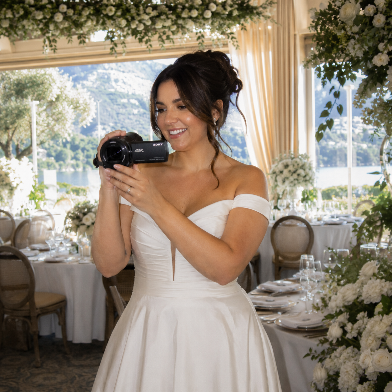 A woman in a wedding dress smiling and holding a camcorder rental in a decorated event space with tables and a scenic view of water and mountains outside.