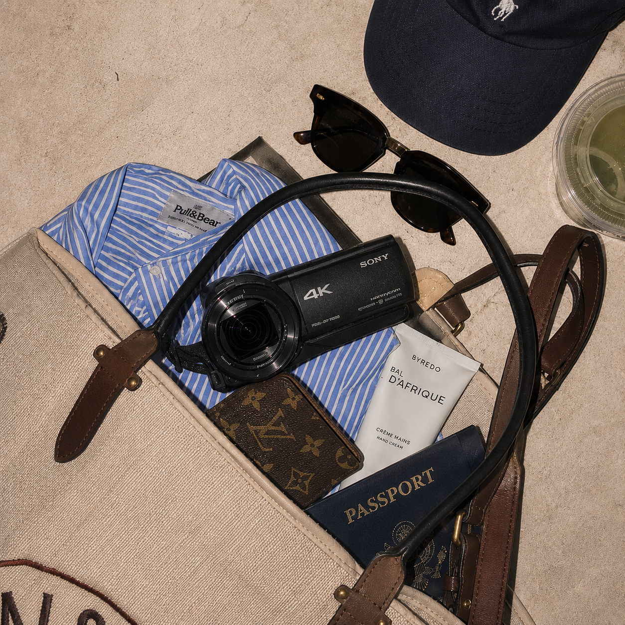 Travel essentials including a camcorder rental from film yours, passport, sunglasses, a striped shirt, a Louis Vuitton wallet, a hand cream, a cap, and a drink in a clear cup in a beige bag and on a beige surface.