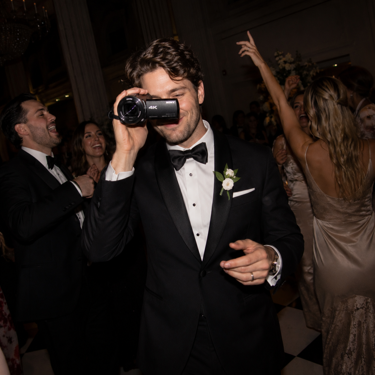 A man in a tuxedo with a bow tie and boutonniere is smiling and holding a camcorder rental at a lively wedding reception or celebration, surrounded by people dancing and enjoying themselves.