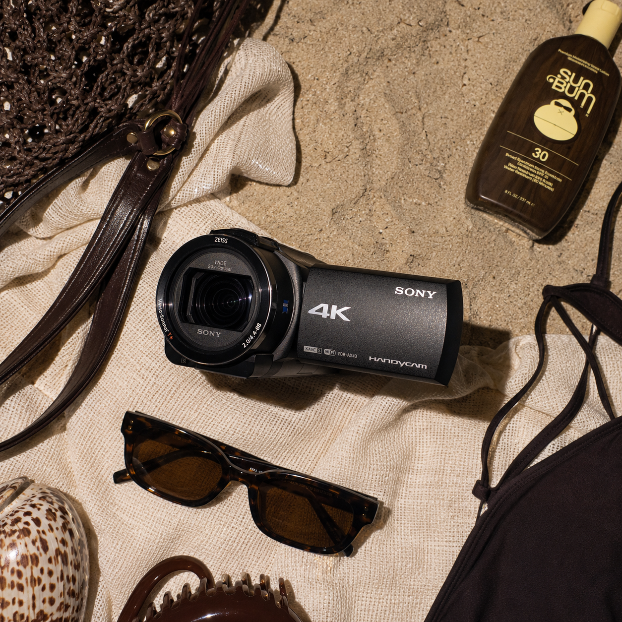 assortment of summer beach accessories on sand, including a 4k camcorder rental, sunglasses, a woven straw bag, sunscreen lotion, a shell, hair clip, and a face mask.