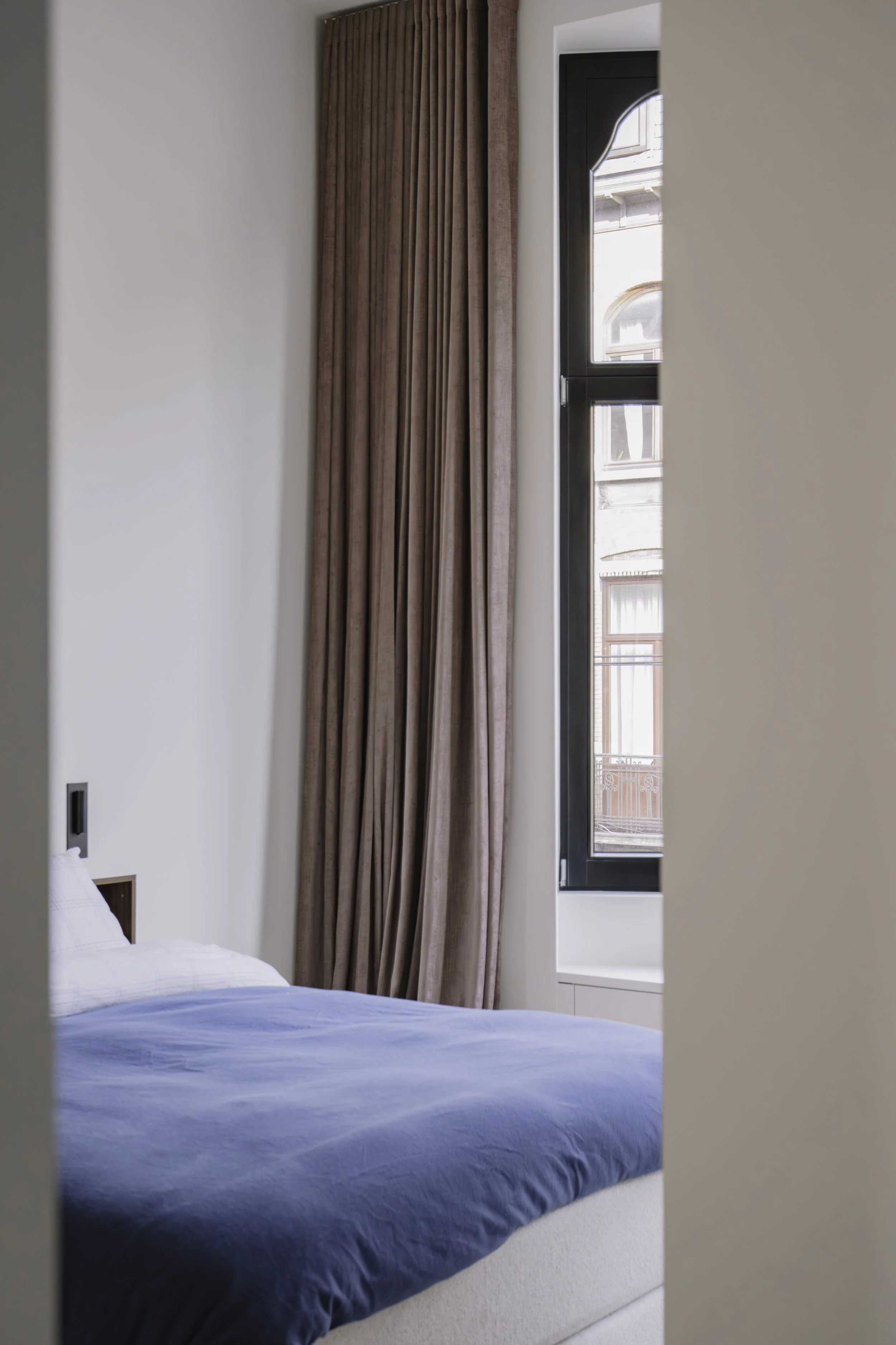 Sfeervolle interieurfotografie van een slaapkamer met blauwe sprei, hoge ramen en bruine gordijnen.