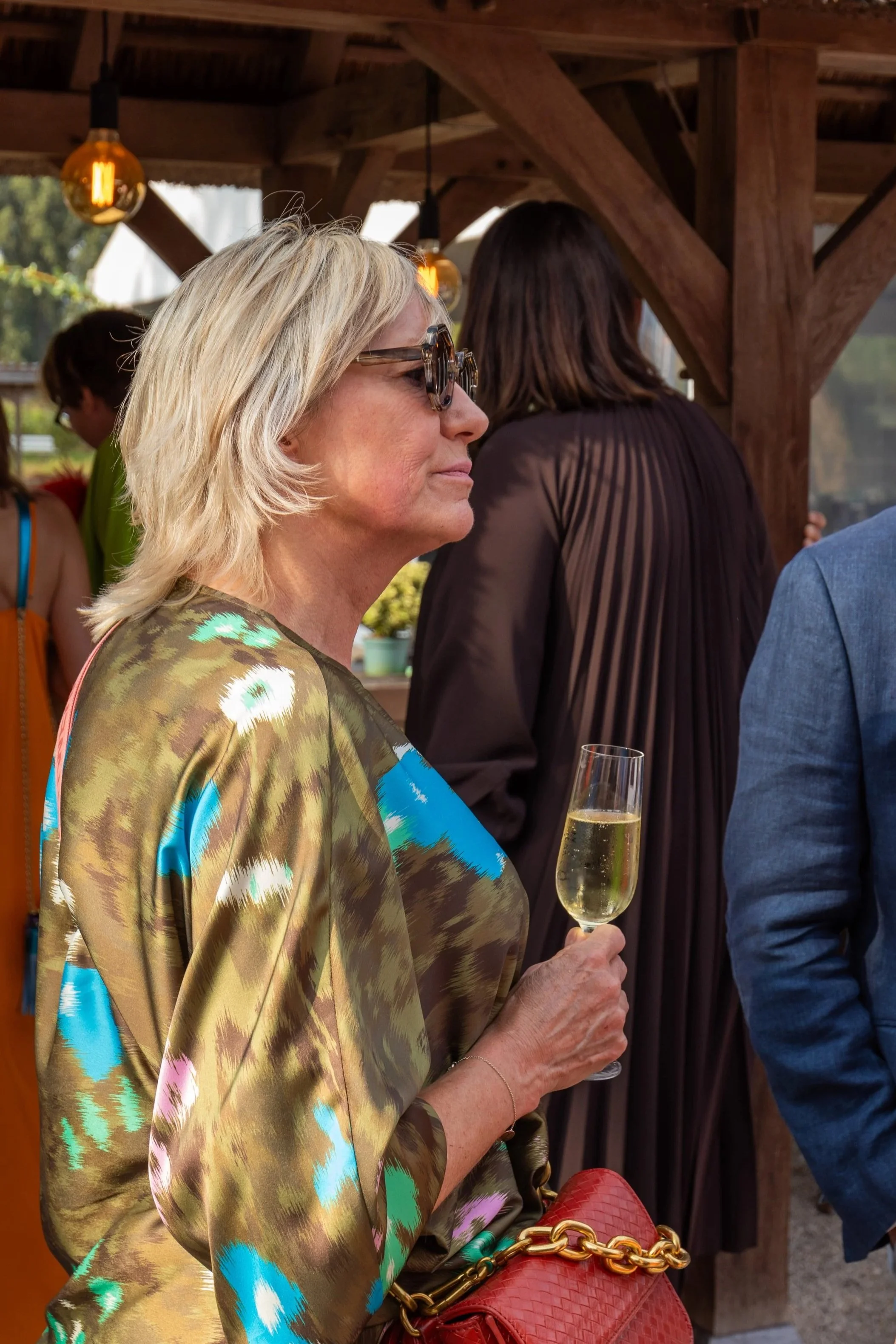 Vrouw met zonnebril en kleurrijke jurk geniet van champagne tijdens het familiefeest.