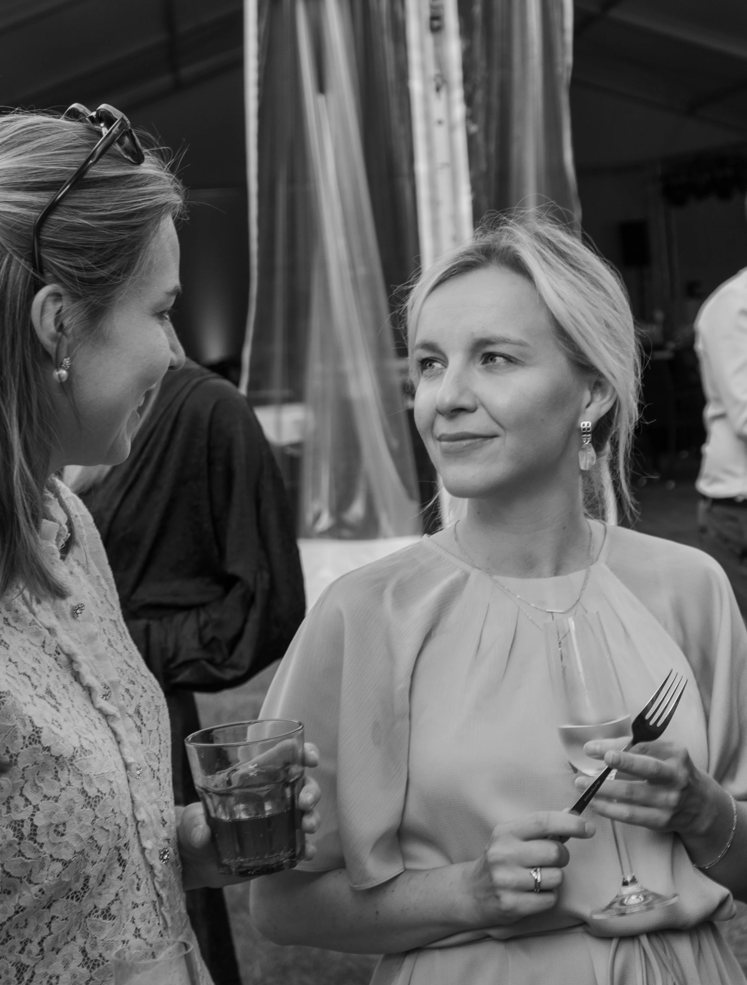 Zwart-witfoto van twee vrouwen in gesprek met drankjes tijdens een evenement.