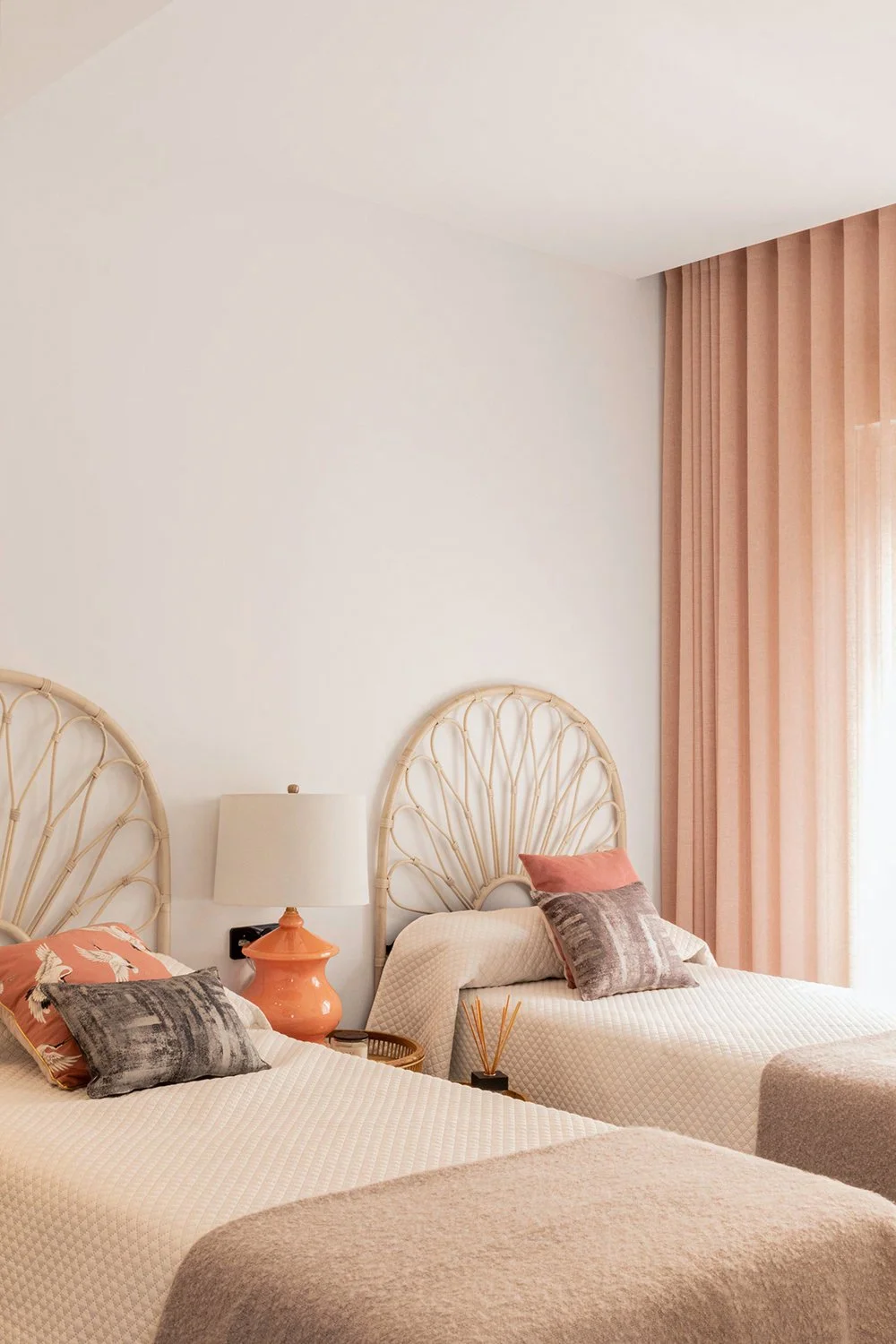 Sfeervolle slaapkamer met twee bedden, rotan hoofdborden en zachtroze gordijnen.