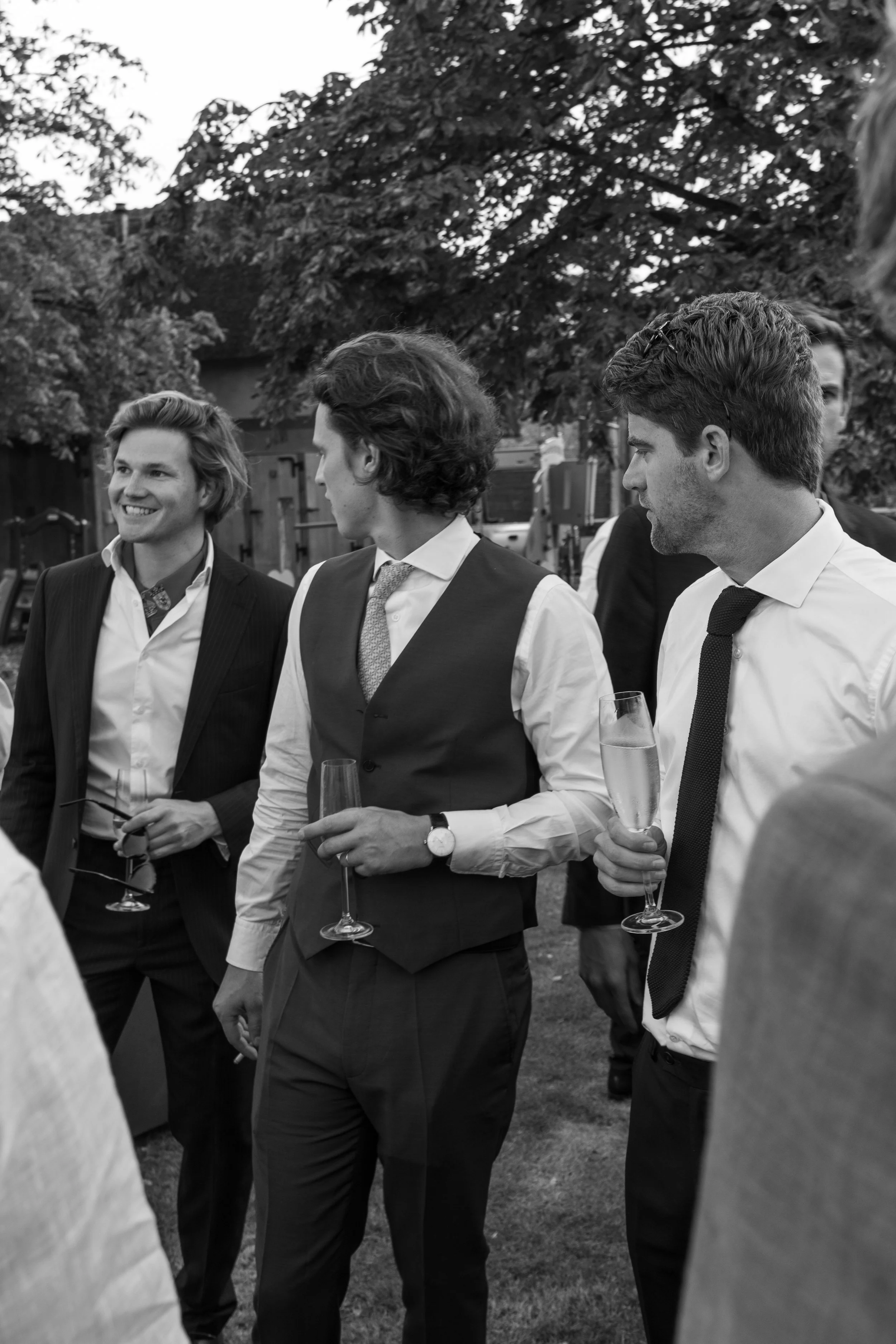 Zwart-witbeeld van stijlvol geklede mannen met champagneglazen tijdens een buitenevenement.