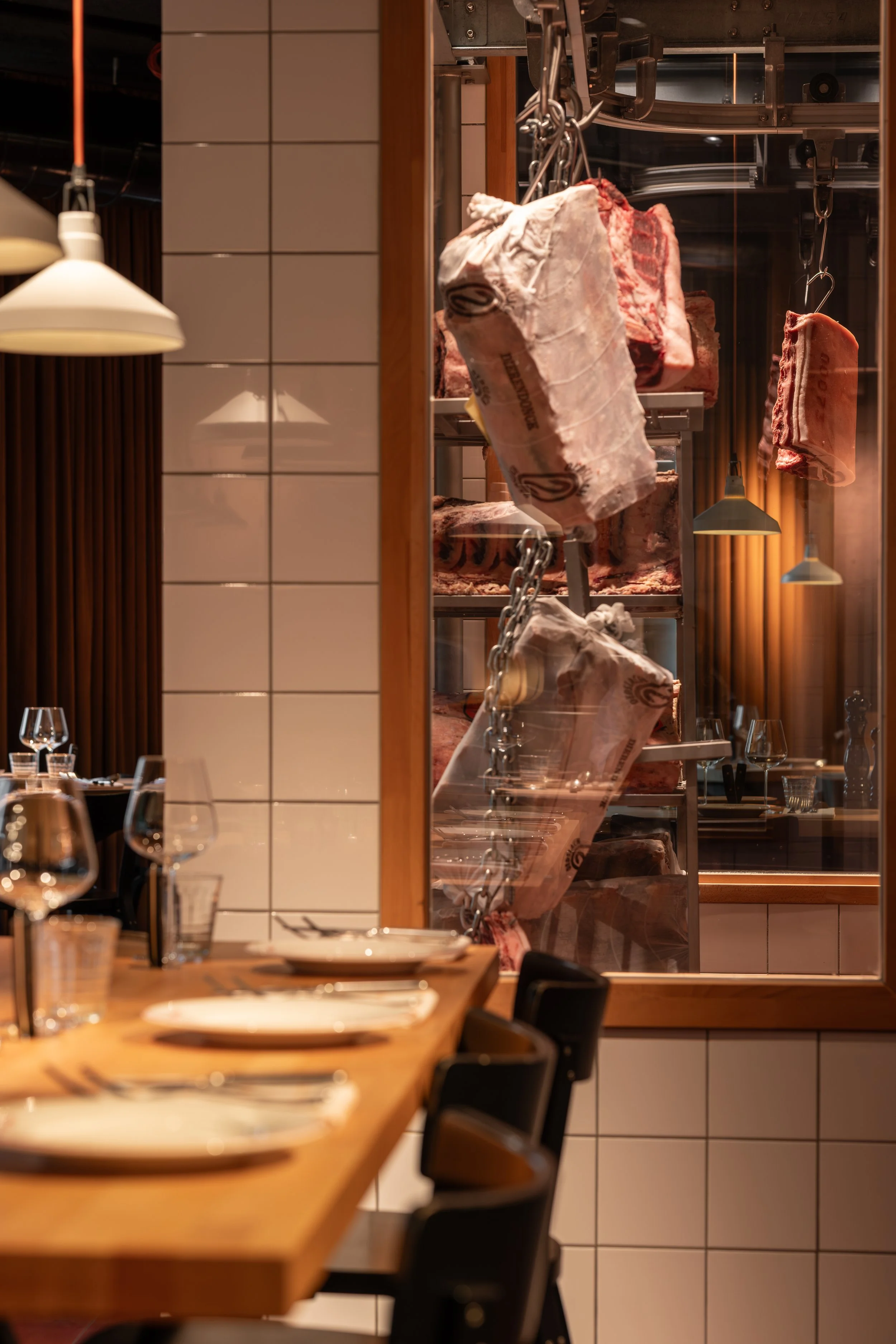 Restaurantinterieur met zicht op gerijpt vlees achter glas en gedekte tafels op de voorgrond.