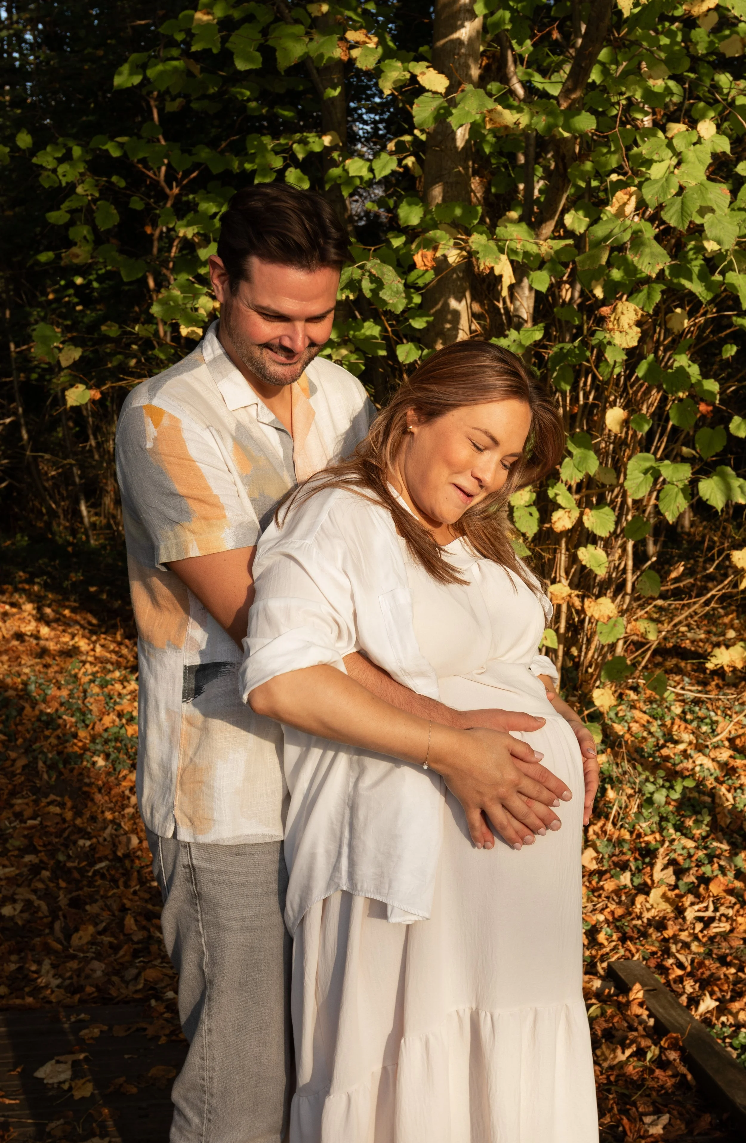 Man omhelst zijn zwangere partner en houdt liefdevol haar buik vast tijdens een herfstshoot.
