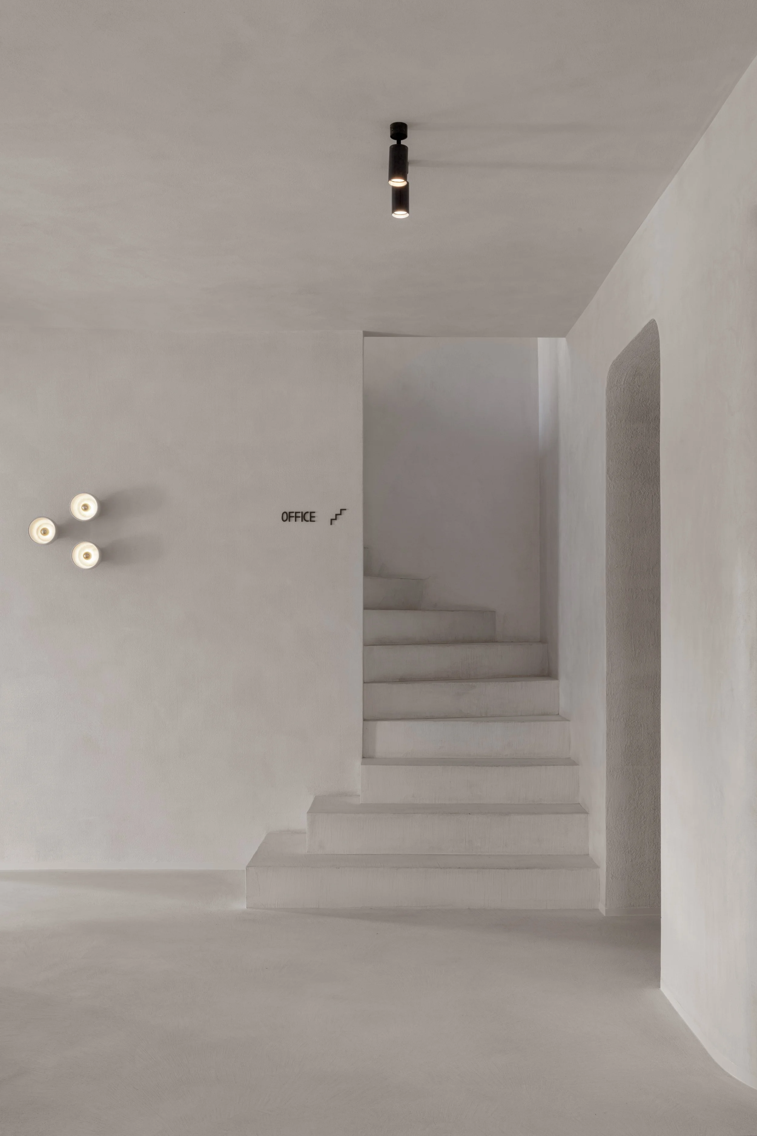 Minimalistische witte inkomhal met trappen, ronde doorgangen, wandverlichting en bewegwijzering naar het kantoor.