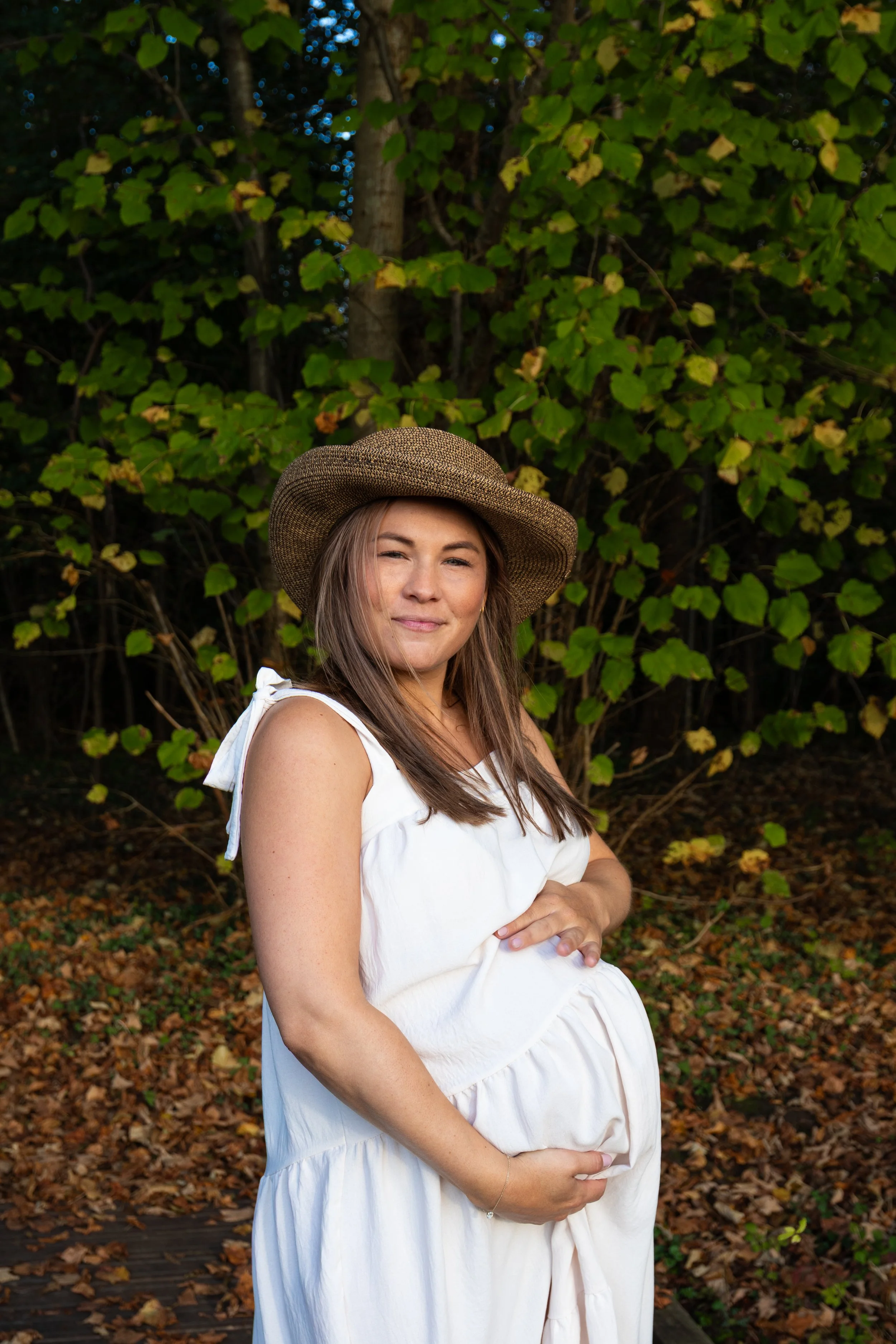 Zwangere vrouw in een witte jurk en strohoed poseert in de natuur.